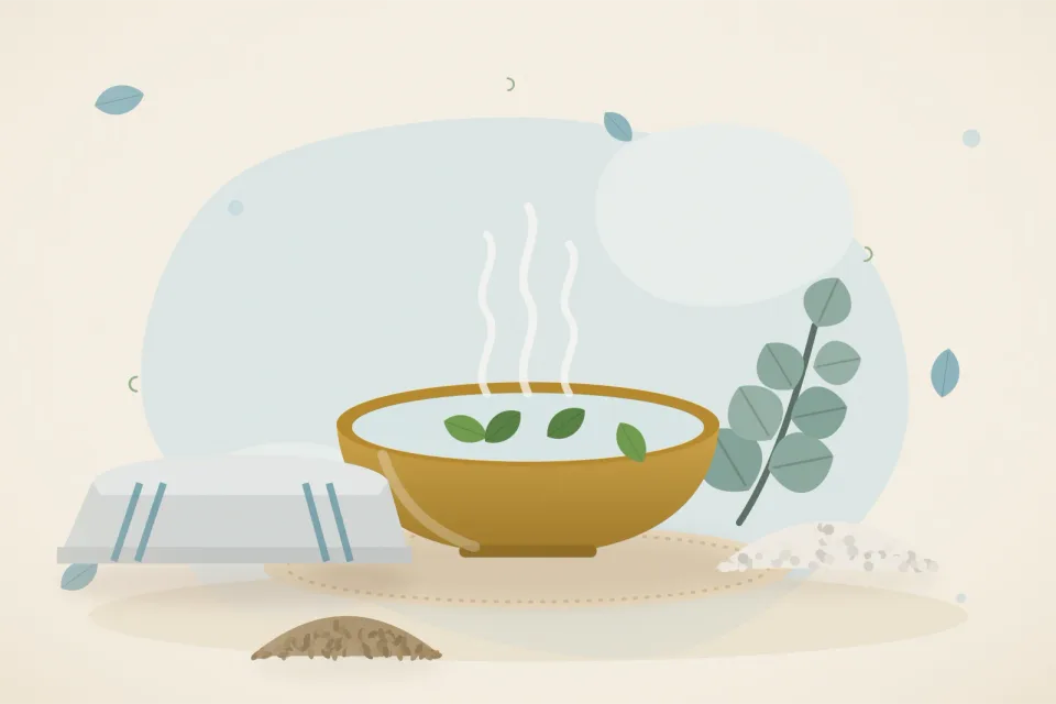 A steaming brass bowl of water with eucalyptus leaves, ajwain seeds, a small bowl of salt and a folded towel on an icy-blue background
