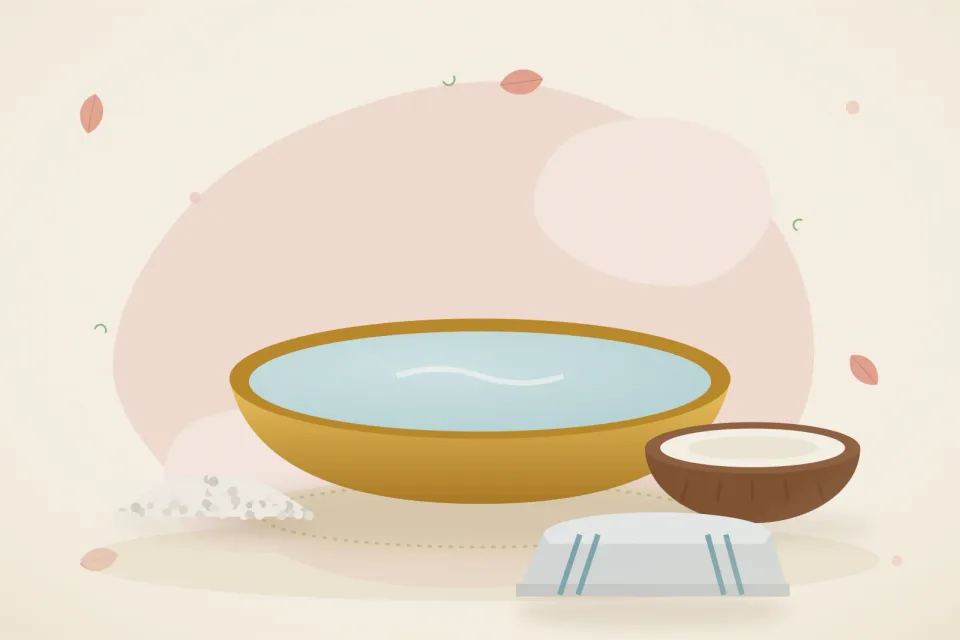 A brass basin of warm water, a small bowl of salt, half a coconut with coconut oil and a folded towel on a rose-pink background